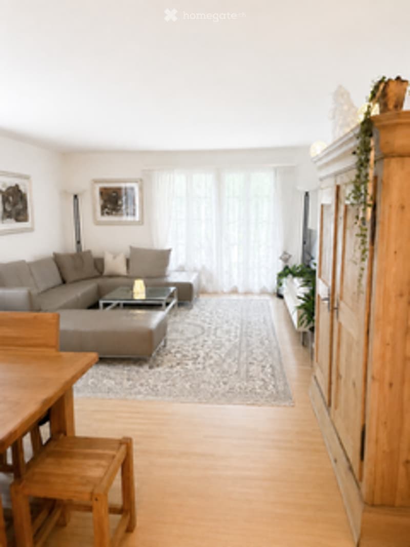 Bright living room with large windows, grey sectional sofa, coffee table, dining table with chairs, floor lamp, and picture frames on the wall.
