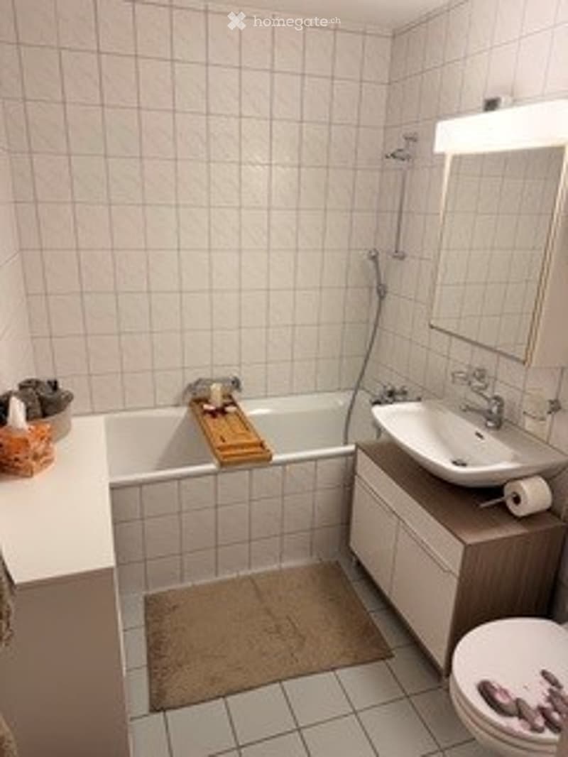 Bathroom with white tiled walls and floor, bathtub, sink, toilet, mirror, and a small cabinet. A towel and a basket are placed on the floor.