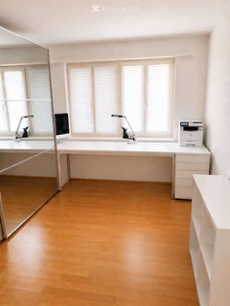 Bright, minimalist room with light wooden flooring, white walls, large windows with blinds, a long desk with chairs, a printer, and a mirror on one wall.