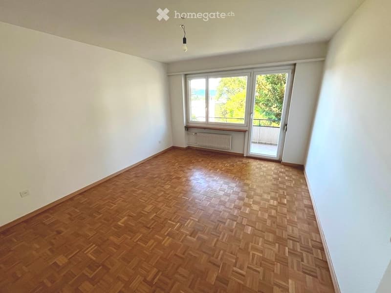 Empty room with wooden parquet flooring, white walls, large window, and sliding glass door leading to a balcony.