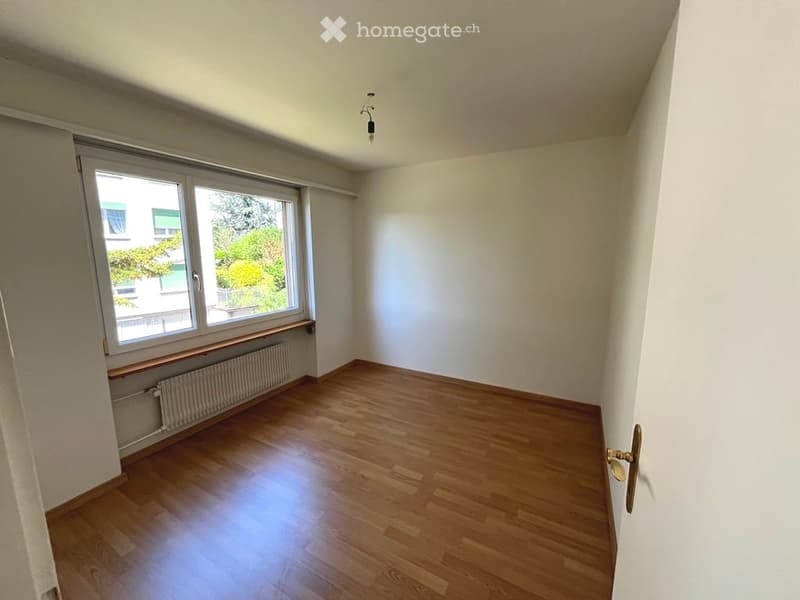 Empty room with wooden flooring, white walls, large window with view, radiator, and a door handle visible.