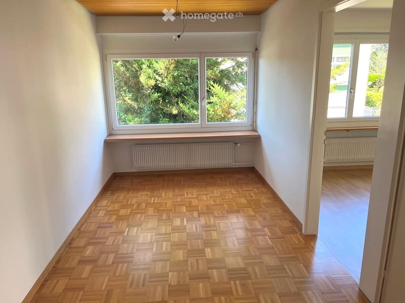 Empty room with wooden parquet flooring, white walls, large window with view of greenery, radiator, and a wooden ceiling.