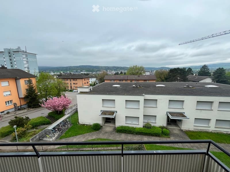 View from a balcony showing a multi-story building with a flat roof, several windows, and surrounding greenery including trees and a small garden area.