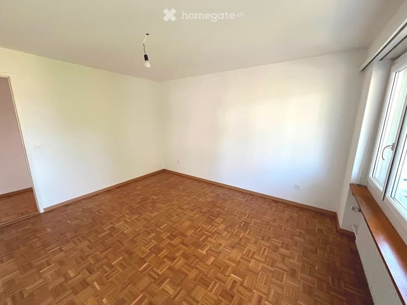 Empty room with wooden herringbone flooring, white walls, and a single light fixture hanging from the ceiling. There is a window with a wooden sill on one wall and a door on another.