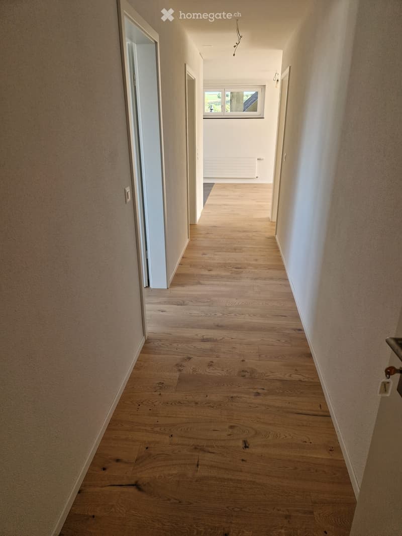 A long, narrow hallway with light wooden flooring and white walls, featuring several doors and a window at the far end.