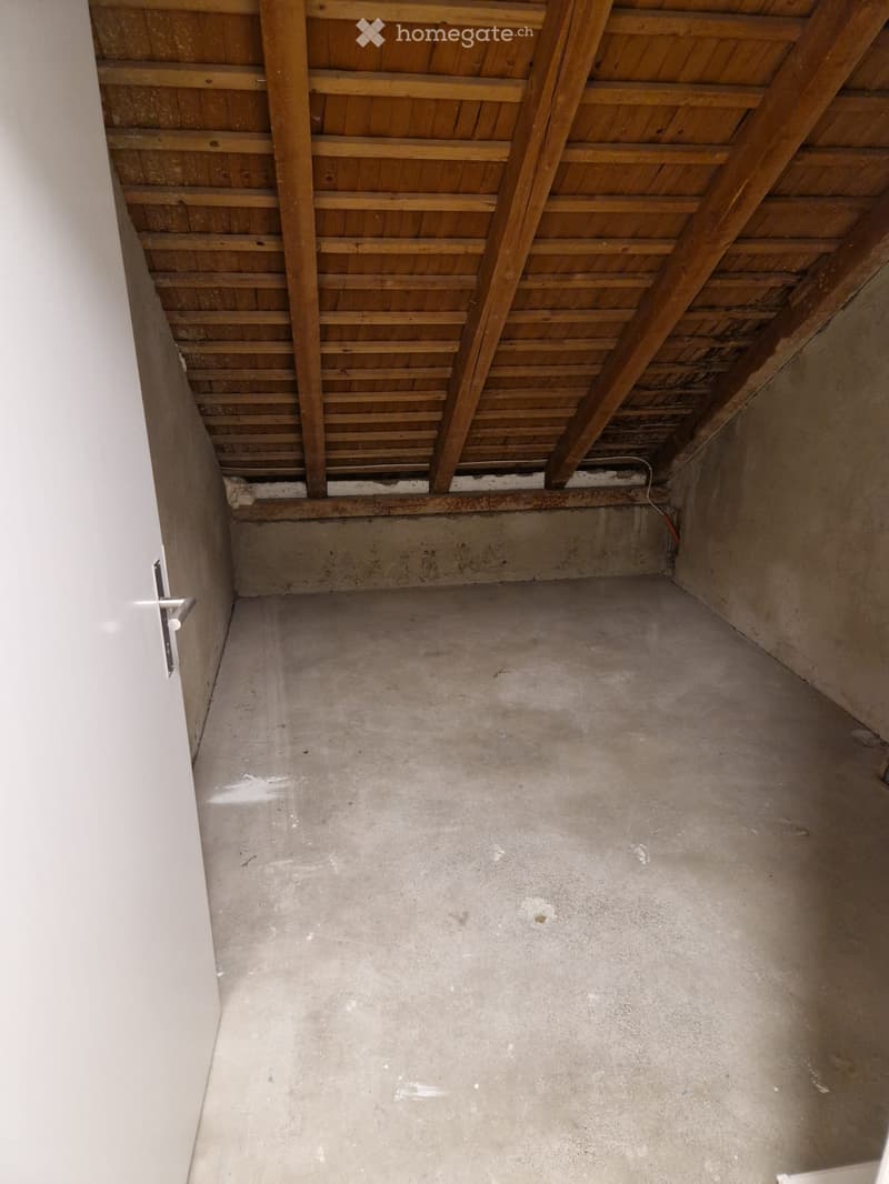 Empty room with concrete floor, exposed wooden ceiling beams, and a single door visible on the left side.