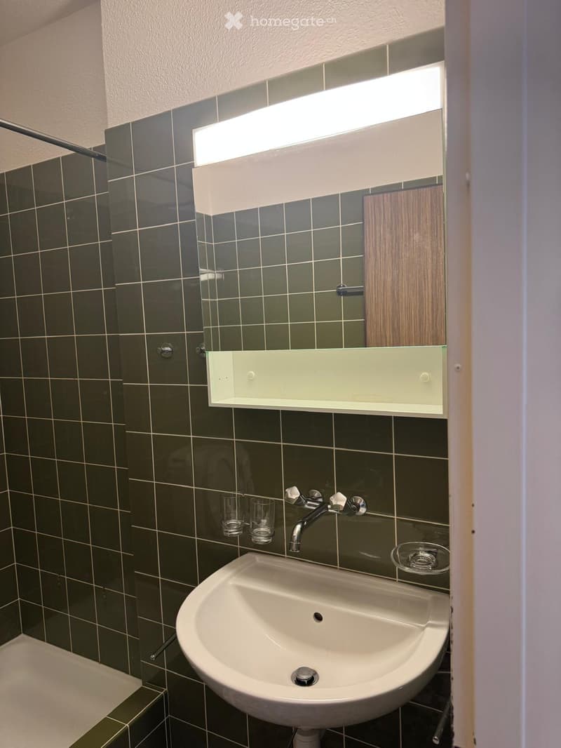 Bathroom with dark green tiled walls, a white sink, a mirror with built-in lighting, and two cups on the counter.