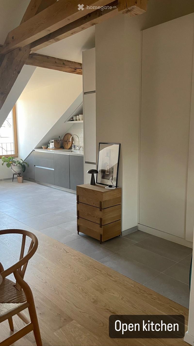 Open kitchen with wooden floors, grey cabinets, a wooden chair, a small wooden cabinet with drawers, a mirror on top, and a small plant in a pot near the window.