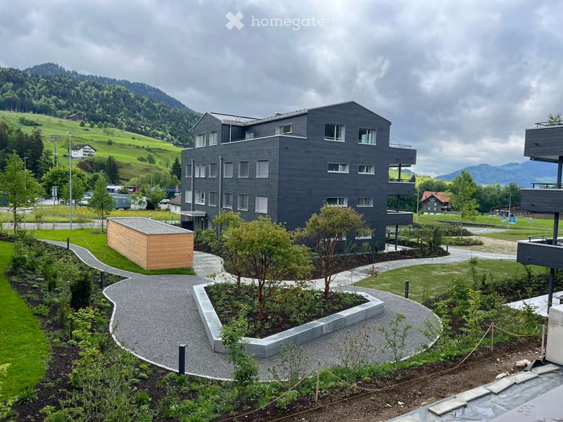 Modern two-story building with grey facade and multiple windows, surrounded by landscaped garden with gravel pathways, small trees, and shrubs. The building features a balcony on the upper level and is situated in a green, hilly area with distant mountain