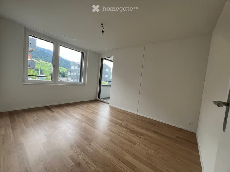 Empty room with light wooden flooring, white walls, and large windows offering a scenic view of a green landscape and neighboring buildings.