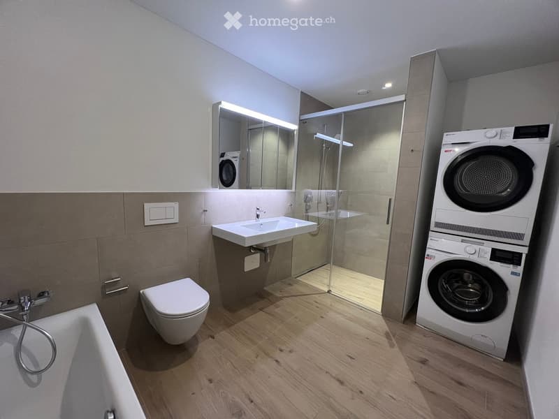 Modern bathroom with a toilet, sink, bathtub, shower, and a washing machine. Features include a large mirror, wooden flooring, and sleek design.