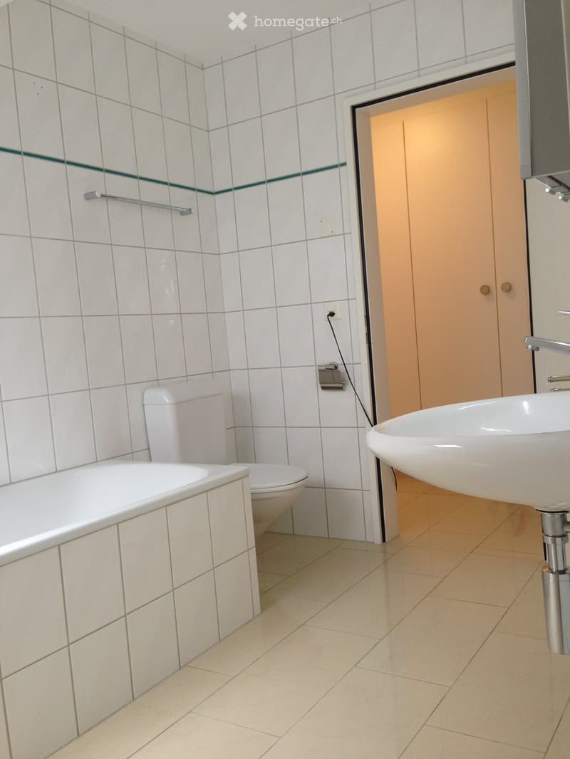 Modern bathroom with white tiled walls, a bathtub, toilet, and a sink with a chrome faucet. The floor is tiled in light beige, and there is a towel rack on the wall.