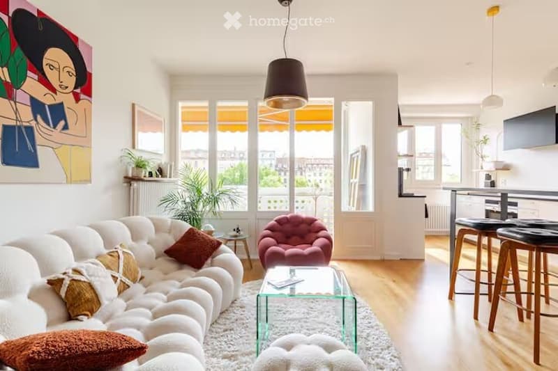 Modern living room with white walls, large windows, a white sectional sofa, a pink armchair, a glass coffee table, and a small round stool. The room features pendant lamps, a painting, and a small plant on the windowsill.