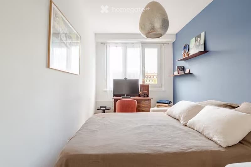 A modern bedroom with a large bed, a wooden desk with a chair, a television, shelves with decorative items, and a large window with white curtains.