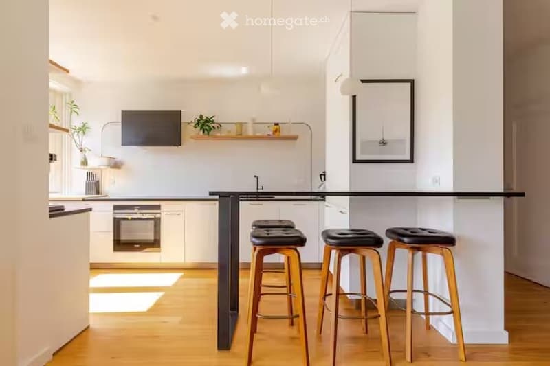 Modern kitchen with white cabinets, wooden floor, island with black countertop, three wooden stools, sink, oven, screen, mirror, and plant.