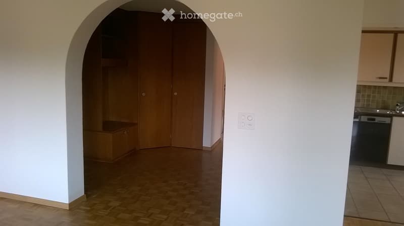 Empty hallway with wooden floor, white walls, and an arched doorway leading to a closet area. The adjacent room shows a kitchen with cabinets and a dishwasher.