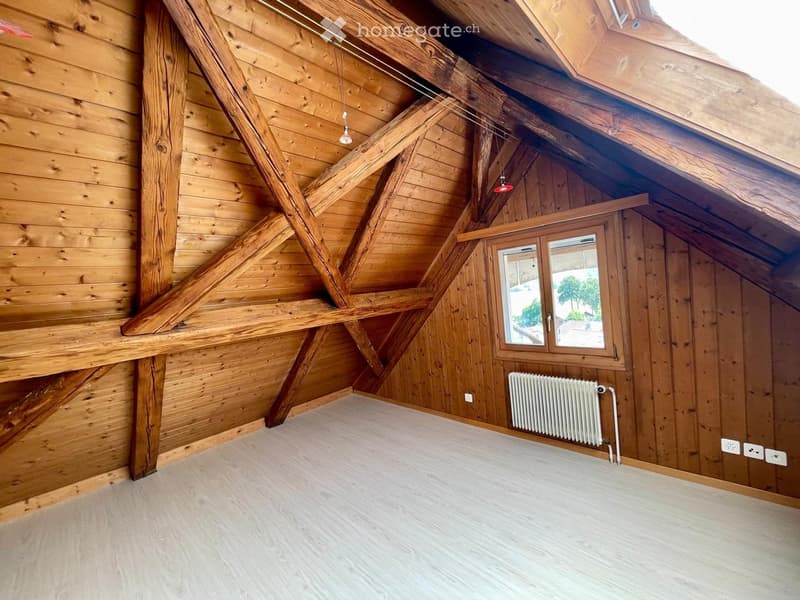 Empty attic room with wooden walls, exposed wooden beams, a skylight window, and a radiator. The floor is covered with light-colored carpeting.