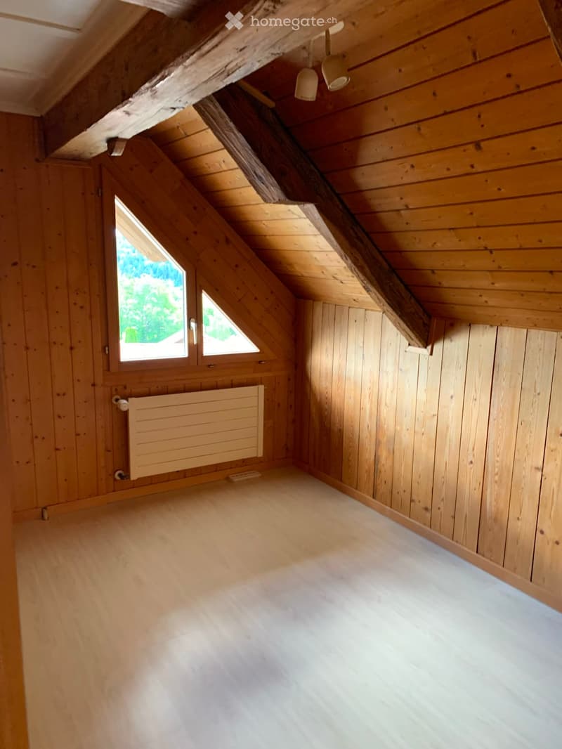 Empty attic room with wooden walls, wooden floor, and skylight windows. Exposed wooden beams on the ceiling and a radiator on the wall.