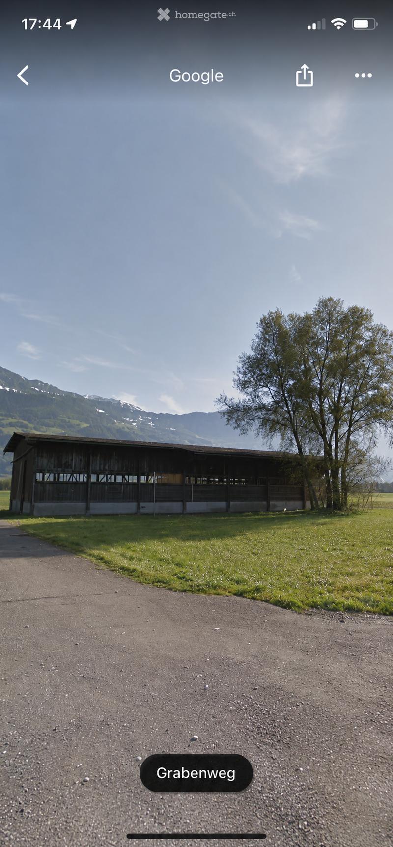 A wooden barn or stable structure with a flat roof, located in a grassy field with trees and mountains in the background.