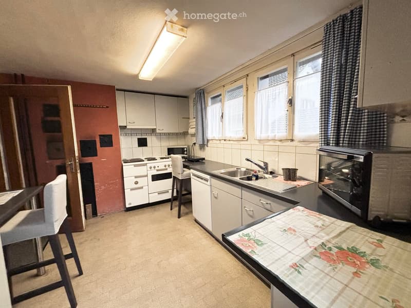 A compact kitchen with white cabinets, a stove, sink, microwave, and a small table with a floral tablecloth. The space features checkered curtains and a single chair.