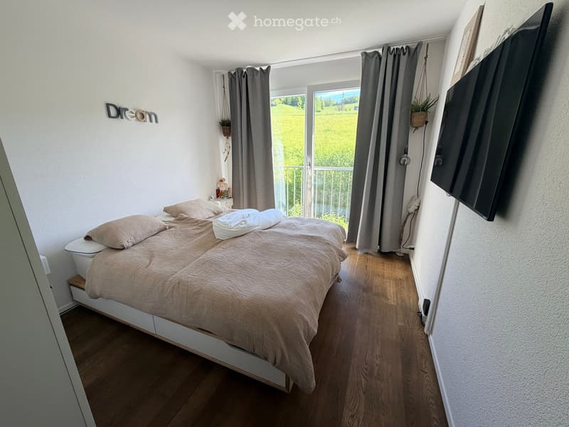 Cozy bedroom with a wooden floor, a large bed with beige bedding, a nightstand, a television mounted on the wall, and a view of a green field through large windows.