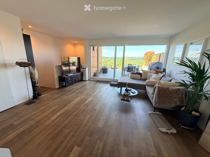 Modern living room with wooden flooring, large windows offering a scenic view, a grey sectional sofa, a round coffee table, a television, and a side table with a floor lamp.