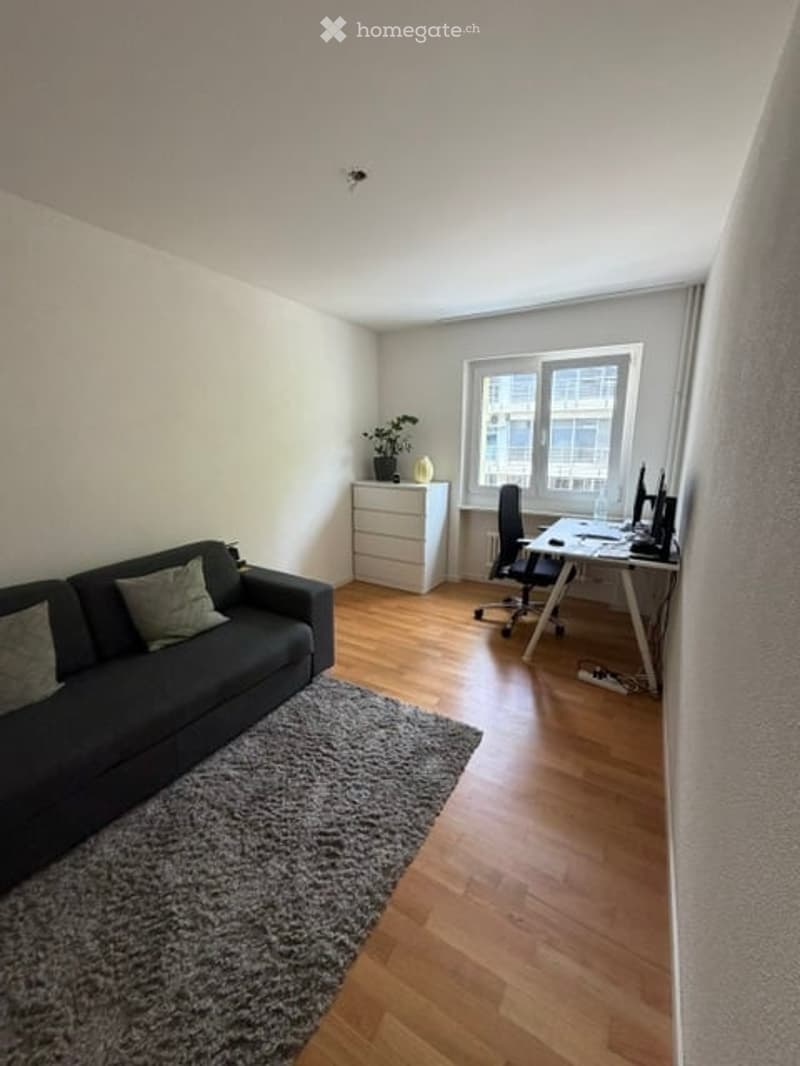 A modern, well-lit living room with a dark grey sofa, a coffee table, a desk with a computer, a chair, a small cabinet with a plant, and a large window with curtains.