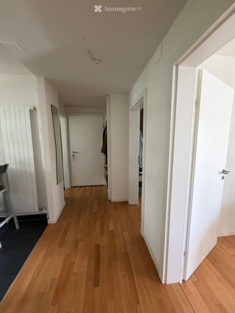 A clean, well-lit hallway with light wooden flooring, white walls, and several doors leading to different rooms.