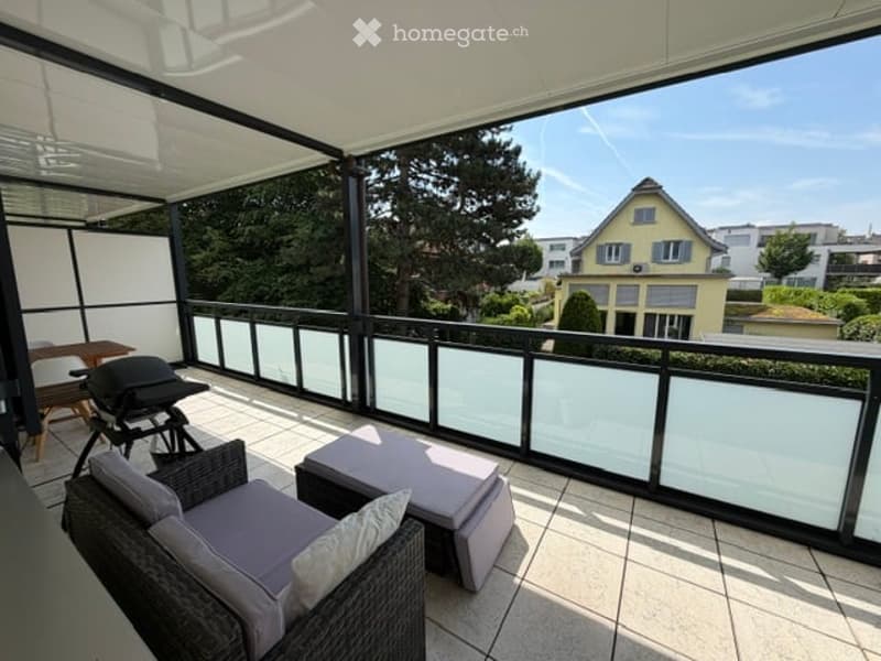 Balcony with modern furniture, including a wicker sofa, ottoman, and a small table, overlooking a residential area with trees and houses.