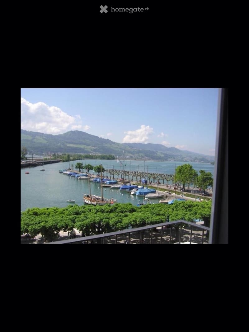 Balcony overlooking a waterfront with boats, trees, and distant mountains under a clear sky.