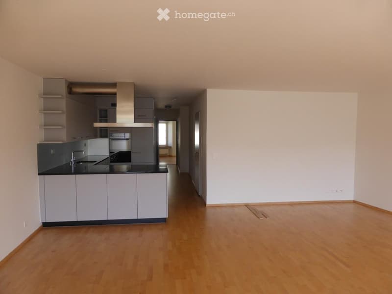 Empty room with light wooden flooring, white walls, and a small kitchen area with cabinets and a sink visible in the background.