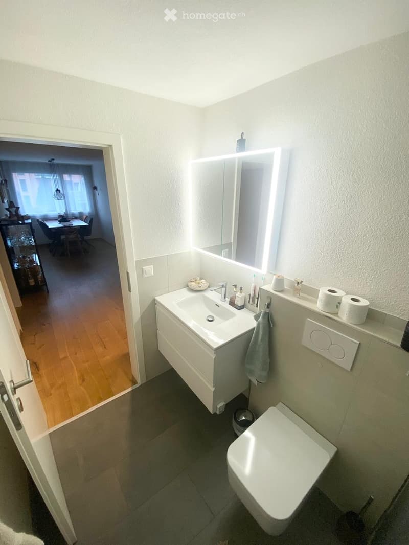 Modern bathroom with a white sink, mirror, toilet, and various toiletries. The room features light-colored tiles and a wooden floor visible through an open doorway.