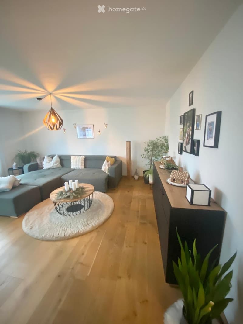 Modern living room with light wooden flooring, grey sectional sofa, round coffee table with candles, pendant light fixture, side table, and decorative plants.