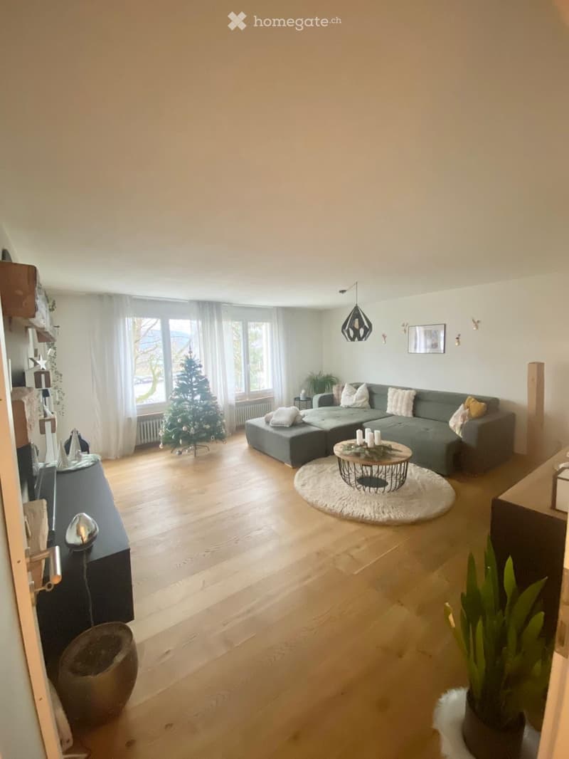 Modern living room with light wooden flooring, large windows, grey sectional sofa, coffee table with a round rug, and a small Christmas tree.