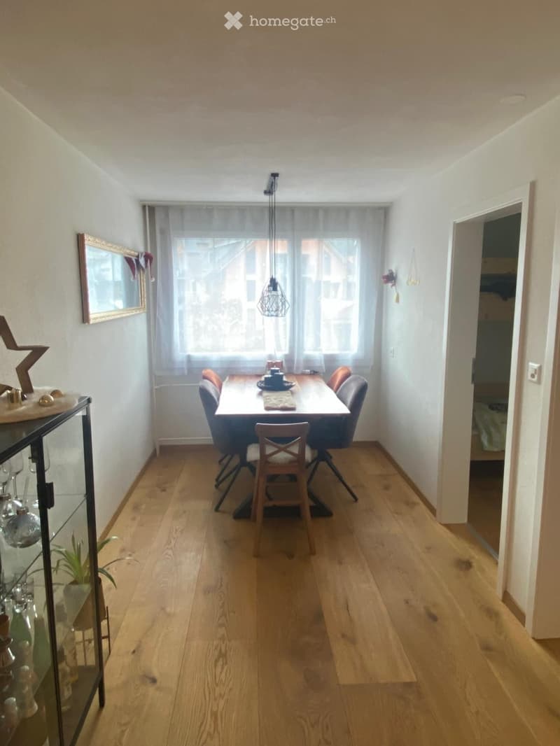 Dining room with wooden floor, large window with sheer curtains, dining table with chairs, pendant light, cabinet with glassware, and a small plant.