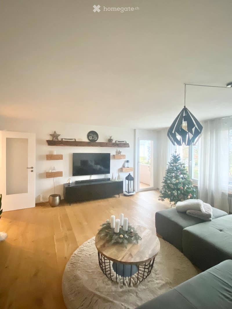Modern living room with wooden flooring, large windows, a grey sofa, a round coffee table with a decorative arrangement, a television on a dark cabinet, and a Christmas tree near the window.
