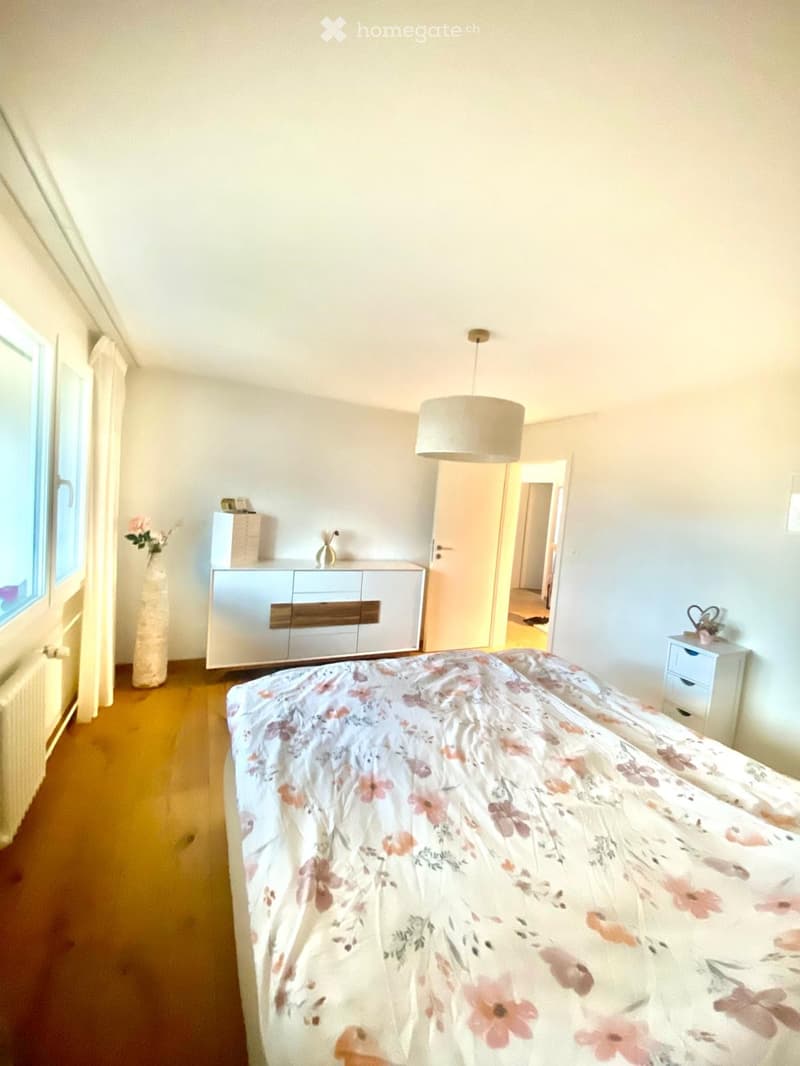 Bright bedroom with wooden flooring, large window, bed with floral duvet, mirror cabinet, vase, and nightstand.