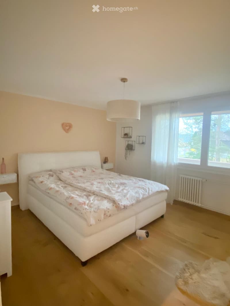 A well-lit bedroom with a white bed, floral bedding, a nightstand, a hanging lamp, and a large window with sheer curtains.