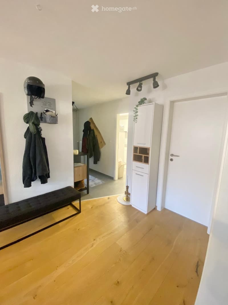 A well-lit hallway with wooden flooring, a bench, a coat rack with coats and a helmet, and a white cabinet with drawers and shelves.