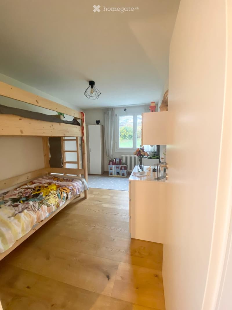 Bright bedroom with wooden bunk bed, wardrobe, small table with vase, and toy house on floor.