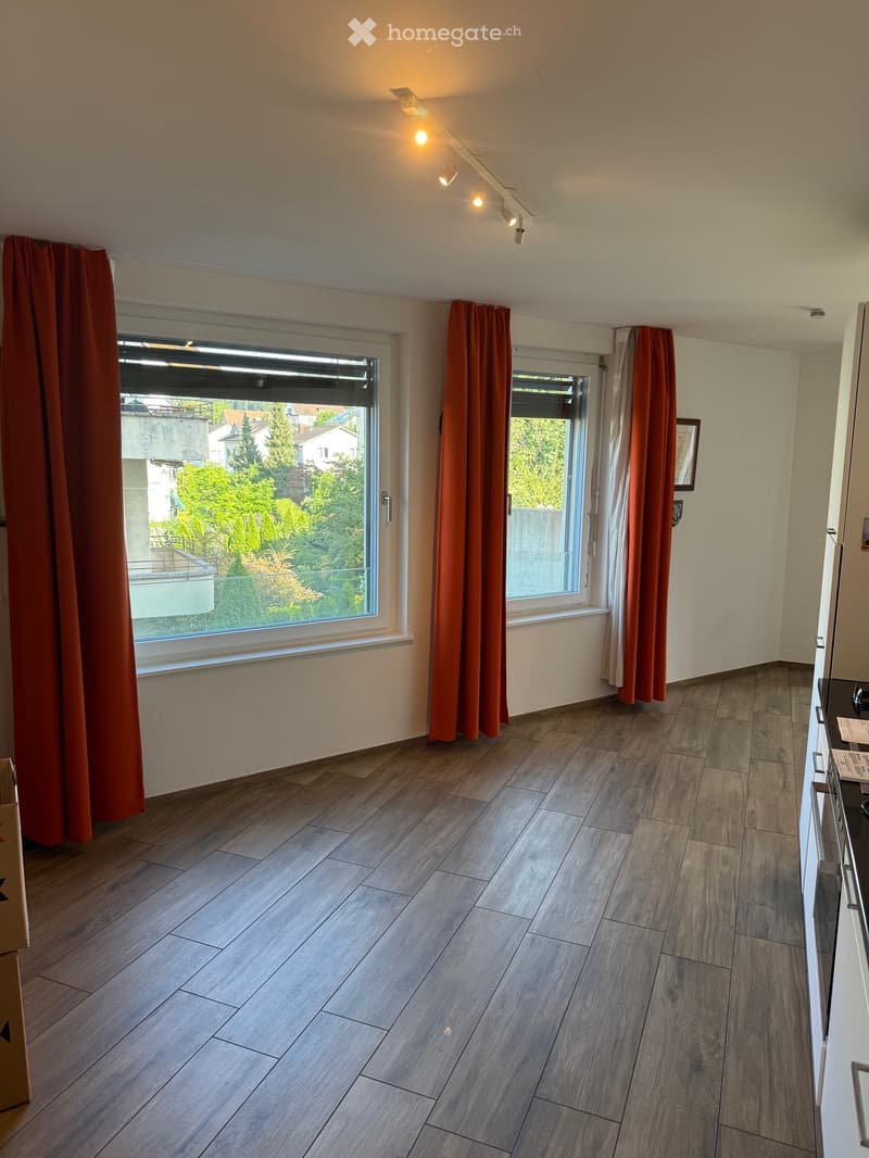 Empty room with wooden flooring, large windows with orange curtains, and track lighting on the ceiling.