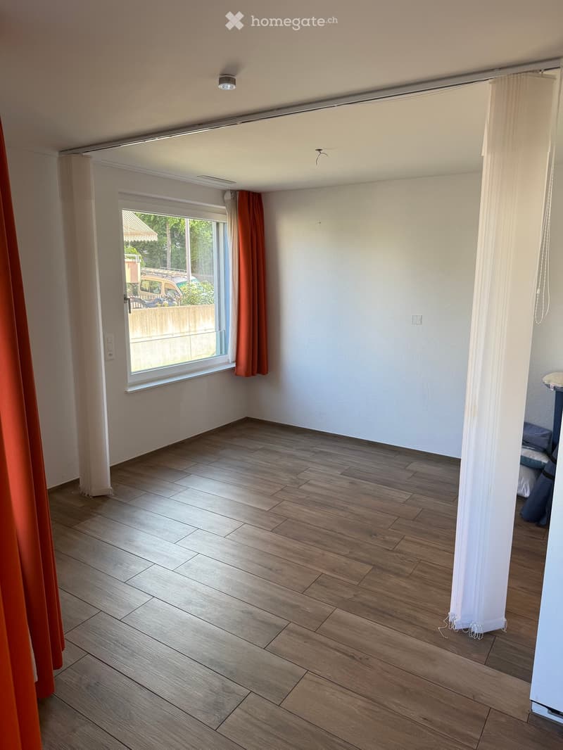 Empty room with wooden flooring, white walls, and large window with orange curtains. No furniture or equipment visible.