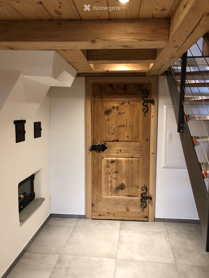 A modern hallway with a wooden door, exposed wooden beams on the ceiling, and a built-in fireplace on the left side.