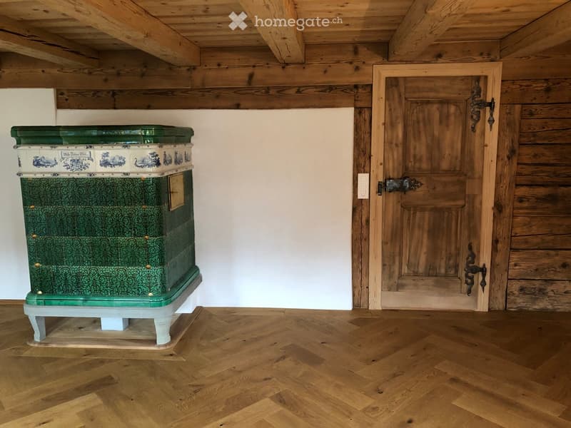 Empty room with wooden floor, exposed wooden beams on the ceiling, and a wooden door with metal hardware. A decorative green and white stove is present.
