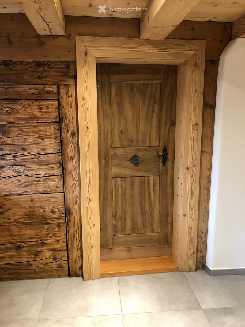 Wooden door with ornate handle and lock, set in a wooden-framed opening with a raised wooden step, adjacent to a wooden wall panel and tiled floor.