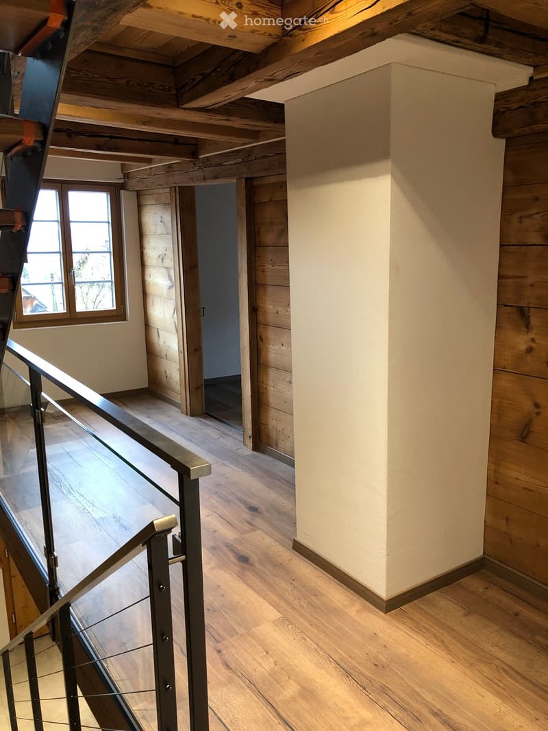 Staircase with wooden floors, metal railing, and exposed wooden beams on the ceiling. The walls are a mix of light plaster and wooden paneling, with a window providing natural light.