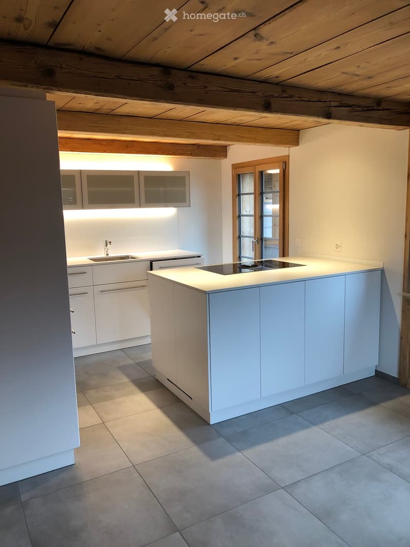 Modern kitchen with white cabinets, a central island, and built-in appliances, including a sink, faucet, and stove. The space features exposed wooden beams on the ceiling and a large window providing natural light.
