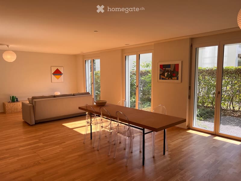 Bright living and dining area with wooden flooring, large windows, a sofa, a dining table with chairs, and decorative artwork on the walls.