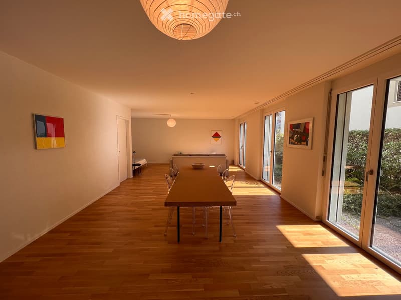 Bright, open dining area with wooden flooring, large dining table surrounded by chairs, a sofa in the background, and modern artwork on the walls.
