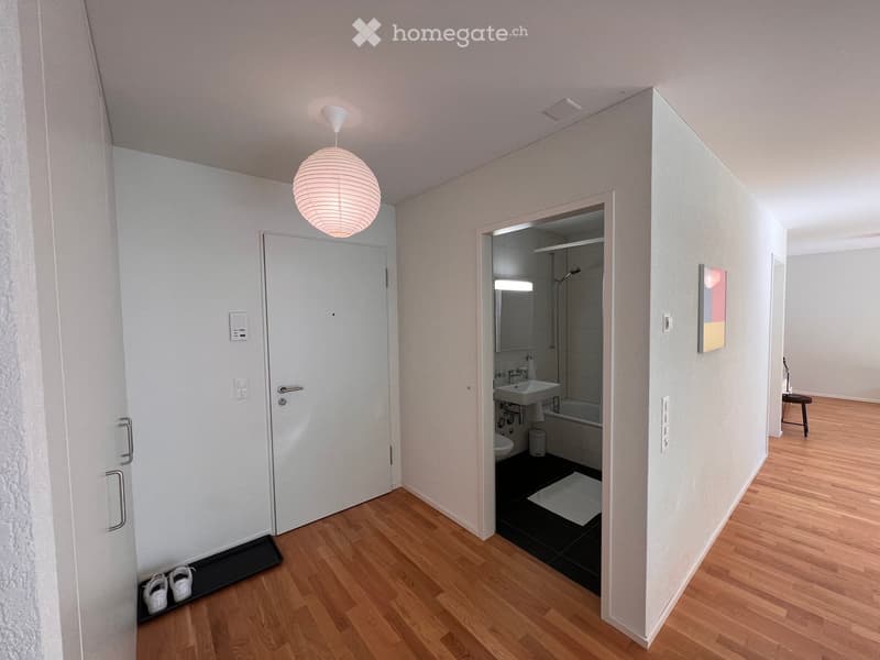 Modern hallway with wooden flooring, white walls, a pendant light fixture, a closed door, and an open door leading to a bathroom with a sink, bathtub, and shower.